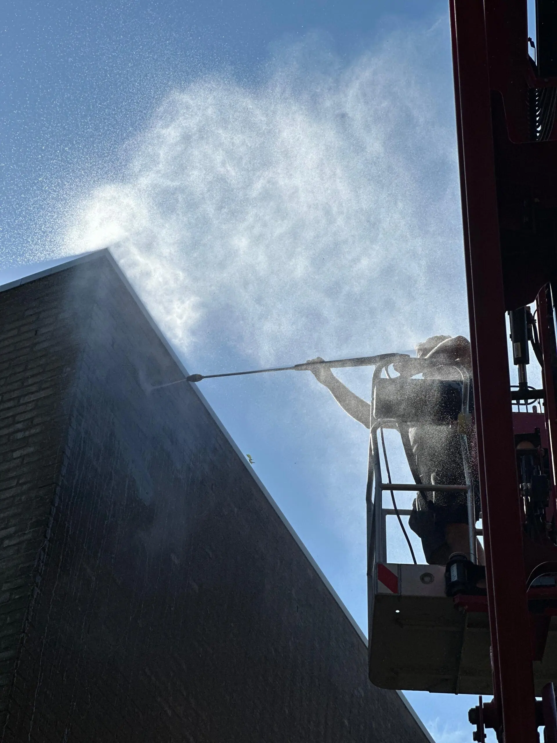 Monteur van LEMA in een hoogwerker reinigt met een hogedrukspuit een bakstenen topgevel, tegen een blauwe lucht.
